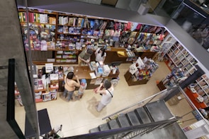 A bookstore with shelves filled with colorful stationery, books, and various items. There are several people interacting at the counter, with an overhead view revealing a bustling and vibrant environment. Items such as notebooks, toys, and art supplies are visible, presenting a lively and organized retail space.