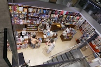 A bookstore with shelves filled with colorful stationery, books, and various items. There are several people interacting at the counter, with an overhead view revealing a bustling and vibrant environment. Items such as notebooks, toys, and art supplies are visible, presenting a lively and organized retail space.