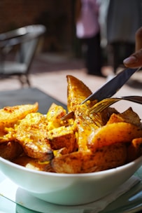 A bowl filled with golden, crispy potato wedges seasoned with herbs and spices. A hand is shown using a fork and knife to cut into one of the wedges, capturing a drizzle of melted cheese.