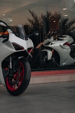 A modern, sleek white motorcycle with red rims is parked in front of a large glass window. Inside the window display, there is another white motorcycle positioned at an angle. The setting appears to be a showroom, with reflective glass and interior lighting casting a sophisticated ambiance.