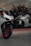A modern, sleek white motorcycle with red rims is parked in front of a large glass window. Inside the window display, there is another white motorcycle positioned at an angle. The setting appears to be a showroom, with reflective glass and interior lighting casting a sophisticated ambiance.