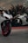 A modern, sleek white motorcycle with red rims is parked in front of a large glass window. Inside the window display, there is another white motorcycle positioned at an angle. The setting appears to be a showroom, with reflective glass and interior lighting casting a sophisticated ambiance.