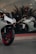 A modern, sleek white motorcycle with red rims is parked in front of a large glass window. Inside the window display, there is another white motorcycle positioned at an angle. The setting appears to be a showroom, with reflective glass and interior lighting casting a sophisticated ambiance.