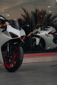 A modern, sleek white motorcycle with red rims is parked in front of a large glass window. Inside the window display, there is another white motorcycle positioned at an angle. The setting appears to be a showroom, with reflective glass and interior lighting casting a sophisticated ambiance.