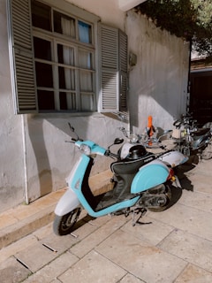 A row of scooters is parked along the side of a building with closed shutters on the windows. The scooters are positioned on a stone-paved surface, and the light blue scooter in the foreground stands out. Sunlight creates shadows against the white wall, adding depth to the scene.