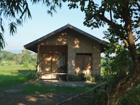 A small, rustic wooden house surrounded by lush greenery and trees. The house is situated in a peaceful rural setting, with sunlight casting shadows through the trees onto the building. The sky is clear, and there are distant hills visible in the background.
