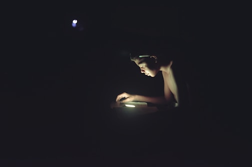 A young person is illuminated by a single light source, using a digital device in a dark environment. The concentrated light highlights their face and hands, creating a strong contrast against the surrounding darkness.