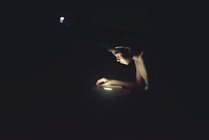 A young person is illuminated by a single light source, using a digital device in a dark environment. The concentrated light highlights their face and hands, creating a strong contrast against the surrounding darkness.