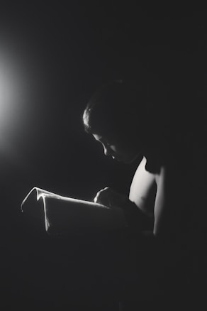 A young person is reading a book in a dimly lit environment, with light casting dramatic shadows and highlighting their focused expression.