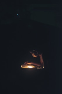 A person is intently reading a book in the dark, illuminated by a warm light source coming from the book itself, creating a focused and intimate atmosphere.