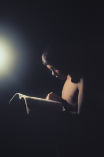 A shadowy figure reading a worn leather-bound book under dim candlelight.