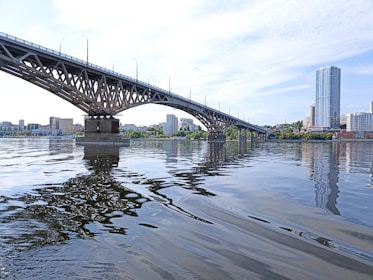 A large bridge extends across a calm body of water, connecting two urban areas. The water reflects the structure and nearby buildings, including a prominent tall skyscraper. Clouds scatter across the bright sky, adding depth to the scene.