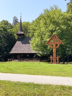 A quaint wooden church with a steep, shingled roof and a tall spire is nestled amidst lush greenery. A wooden cross structure with intricate carvings stands in the foreground on a well-maintained lawn, bordered by a pathway. Trees frame the background, creating a peaceful, natural setting under a clear blue sky.