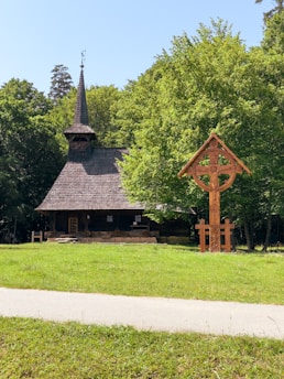 A quaint wooden church with a steep, shingled roof and a tall spire is nestled amidst lush greenery. A wooden cross structure with intricate carvings stands in the foreground on a well-maintained lawn, bordered by a pathway. Trees frame the background, creating a peaceful, natural setting under a clear blue sky.