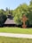 A quaint wooden church with a steep, shingled roof and a tall spire is nestled amidst lush greenery. A wooden cross structure with intricate carvings stands in the foreground on a well-maintained lawn, bordered by a pathway. Trees frame the background, creating a peaceful, natural setting under a clear blue sky.