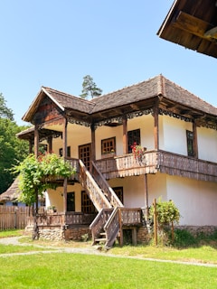 A traditional wooden house with a two-story structure, featuring a rustic staircase leading up to a balcony. The walls are white with exposed timber frames, and there are vibrant red flowers on the balcony railing. The house is surrounded by a lush garden and greenery, with trees visible in the background under a clear blue sky.