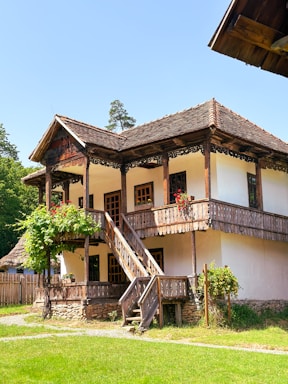 A traditional wooden house with a two-story structure, featuring a rustic staircase leading up to a balcony. The walls are white with exposed timber frames, and there are vibrant red flowers on the balcony railing. The house is surrounded by a lush garden and greenery, with trees visible in the background under a clear blue sky.