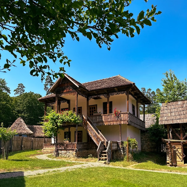 A rustic wooden house surrounded by lush greenery and a clear blue sky. The structure features intricate woodwork and a charming covered balcony adorned with red flowers. A wooden staircase leads up to the balcony, and the house is enclosed by a wooden fence and additional thatched structures.