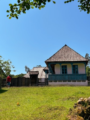 A rustic wooden house with blue accents stands in the middle of a grassy area under a clear blue sky. A person in a red shirt and jeans walks towards the entrance of the property, which is surrounded by a wooden fence. Trees and bushes frame the scene, adding a natural touch.