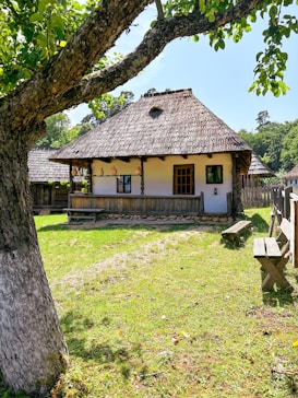 A rustic wooden house with a thatched roof sits in a green grassy area. The structure features a small porch with pottery displayed, and is surrounded by a wooden fence. A tree with green leaves arches over the scene, casting shadows on the grass. The setting appears peaceful and rural.