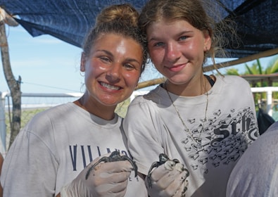 Two smiling individuals are wearing white shirts and are engaged in an outdoor activity. They are holding small sea turtles while wearing protective gloves, with a background of trees and a blue sky.