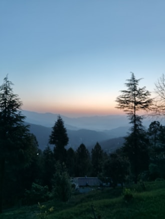 Tall trees frame a serene landscape with rolling hills and a colorful sky at dusk, transitioning from soft orange near the horizon to a deepening blue. The scene is tranquil and expansive, with a small structure nestled among the trees in the lower portion, contributing to a peaceful rural setting.