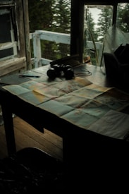 A dimly lit room with a wooden table near a window. Items on the table include a map, a pair of binoculars, and some writing utensils. The window reveals a view of trees, suggesting a setting in a forested area. The scene conveys a sense of solitude and exploration.