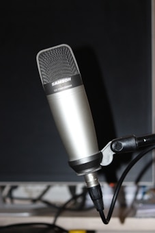A professional studio condenser microphone is mounted on a stand. The microphone has a sleek, metallic body with a mesh grille at the top, displaying a brand name. It is connected via an XLR cable visible in the lower part of the image. The background is dark, highlighting the microphone.