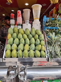 A vibrant fruit juice stall featuring a display of stacked mangoes and pineapples. Several blenders are visible in the background along with a collection of colorful cups. A person stands to the side, ready to serve customers.