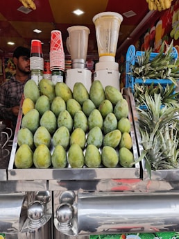 A vibrant fruit juice stall featuring a display of stacked mangoes and pineapples. Several blenders are visible in the background along with a collection of colorful cups. A person stands to the side, ready to serve customers.
