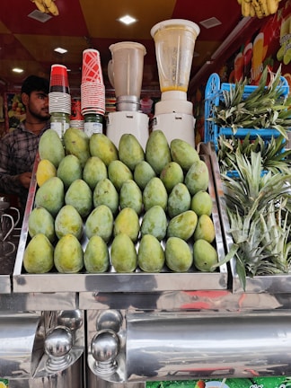 A vibrant fruit juice stall featuring a display of stacked mangoes and pineapples. Several blenders are visible in the background along with a collection of colorful cups. A person stands to the side, ready to serve customers.