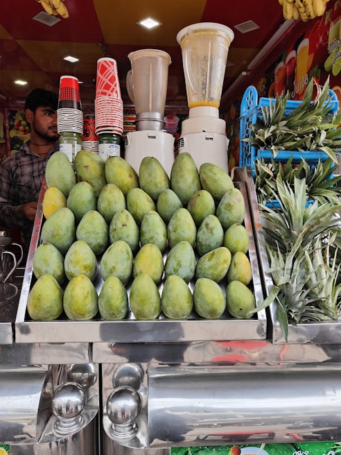 A vibrant fruit juice stall featuring a display of stacked mangoes and pineapples. Several blenders are visible in the background along with a collection of colorful cups. A person stands to the side, ready to serve customers.