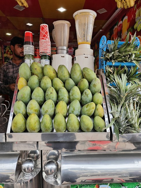 A vibrant fruit juice stall featuring a display of stacked mangoes and pineapples. Several blenders are visible in the background along with a collection of colorful cups. A person stands to the side, ready to serve customers.