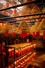 A room filled with large, spiral incense coils hanging from the ceiling, each with a red tag attached. Below, rows of glowing candles or lanterns are aligned on shelves, casting a warm, orange glow throughout the space.