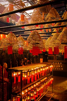 A room filled with large, spiral incense coils hanging from the ceiling, each with a red tag attached. Below, rows of glowing candles or lanterns are aligned on shelves, casting a warm, orange glow throughout the space.