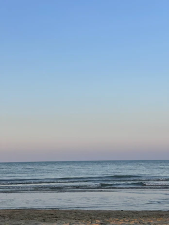A serene seascape showing gentle waves lapping against a quiet beach under a pastel sky.