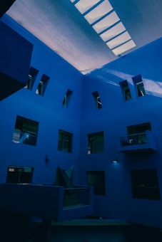 A modern architectural interior with multiple levels, characterized by blue walls and rectangular windows. The design features geometric shapes and asymmetrical elements, including a large skylight on the ceiling that lets in natural light. The area is spacious, and the lighting creates soft shadows on the walls.