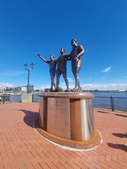 A bronze statue of three rugby players standing arm in arm, located on a brick-paved platform by a waterfront. Each player is depicted in action, with one gesturing forward. In the background, the sky is clear and blue.