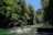 Nine people are rafting on inflatable boats down a clear, rushing river surrounded by lush green forest and towering trees. Sunlight filters through the canopy, highlighting the vibrant greenery and creating dramatic shadows on the water.