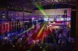 A lively indoor venue filled with people enjoying an event or party, illuminated by colorful lighting effects and beams. Overhead, there's a metal framework supporting numerous spotlights and stage lights. The ambient lighting is accented with vibrant colors, creating an energetic atmosphere.