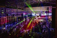 A lively indoor venue filled with people enjoying an event or party, illuminated by colorful lighting effects and beams. Overhead, there's a metal framework supporting numerous spotlights and stage lights. The ambient lighting is accented with vibrant colors, creating an energetic atmosphere.