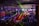 A lively indoor venue filled with people enjoying an event or party, illuminated by colorful lighting effects and beams. Overhead, there's a metal framework supporting numerous spotlights and stage lights. The ambient lighting is accented with vibrant colors, creating an energetic atmosphere.