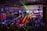 A lively indoor venue filled with people enjoying an event or party, illuminated by colorful lighting effects and beams. Overhead, there's a metal framework supporting numerous spotlights and stage lights. The ambient lighting is accented with vibrant colors, creating an energetic atmosphere.