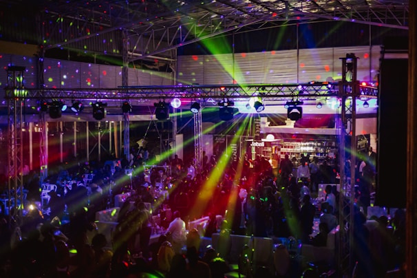 A lively indoor venue filled with people enjoying an event or party, illuminated by colorful lighting effects and beams. Overhead, there's a metal framework supporting numerous spotlights and stage lights. The ambient lighting is accented with vibrant colors, creating an energetic atmosphere.