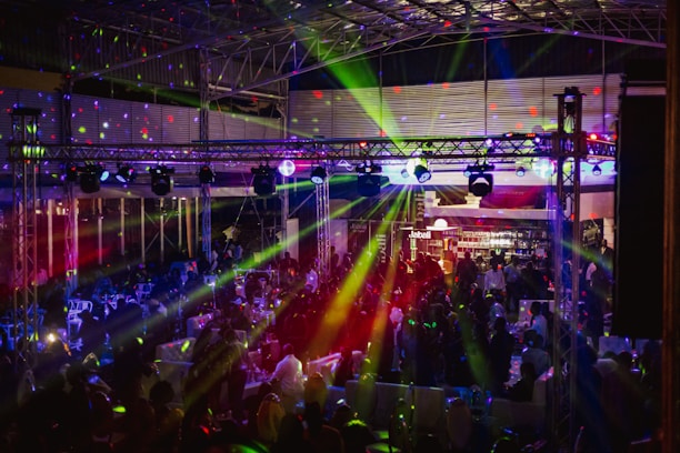 A lively indoor venue filled with people enjoying an event or party, illuminated by colorful lighting effects and beams. Overhead, there's a metal framework supporting numerous spotlights and stage lights. The ambient lighting is accented with vibrant colors, creating an energetic atmosphere.