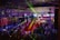 A lively indoor venue filled with people enjoying an event or party, illuminated by colorful lighting effects and beams. Overhead, there's a metal framework supporting numerous spotlights and stage lights. The ambient lighting is accented with vibrant colors, creating an energetic atmosphere.