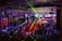 A lively indoor venue filled with people enjoying an event or party, illuminated by colorful lighting effects and beams. Overhead, there's a metal framework supporting numerous spotlights and stage lights. The ambient lighting is accented with vibrant colors, creating an energetic atmosphere.