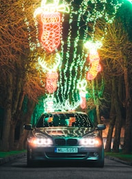 A car with headlights on is parked on a road lined with leafless trees. Overhead, there are vibrant holiday-themed lights in various shapes, including stockings and reindeer, illuminating the scene in bright green and red colors.