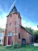 A red brick church with pointed arched windows and a tall steeple with a cross on top. The structure includes white stone accents and a large wooden door. The sky is clear and blue, while some greenery is visible around the building.