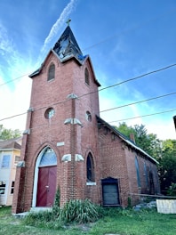 A red brick church with pointed arched windows and a tall steeple with a cross on top. The structure includes white stone accents and a large wooden door. The sky is clear and blue, while some greenery is visible around the building.